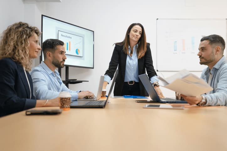 Cuatro profesionales en una sala de reuniones corporativa durante una sesi&oacute;n de estrategia. Una mujer vestida con chaqueta negra est&aacute; de pie en el centro de la mesa liderando la conversaci&oacute;n, mientras tres colegas (dos hombres y una mujer) la escuchan con atenci&oacute;n y revisan documentos impresos y ordenadores port&aacute;tiles. Al fondo, una gran pantalla digital muestra un gr&aacute;fico de barras y un diagrama de sectores bajo el t&iacute;tulo "Resultados", junto a una pizarra con anotaciones estrat&eacute;gicas.